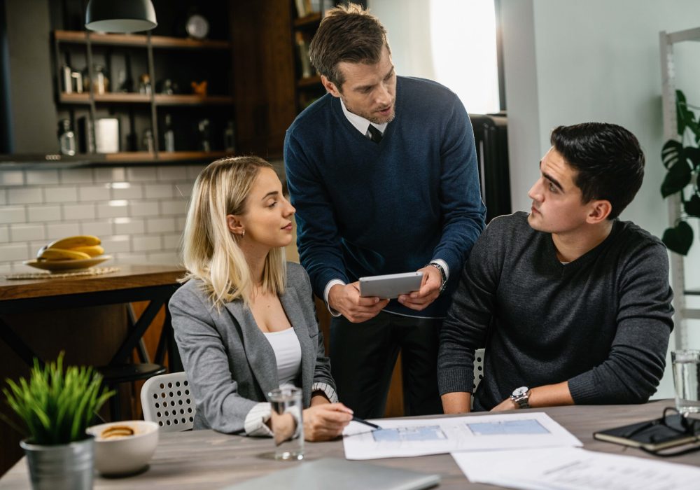 Rear estate agent and young couple going through blueprints and using digital tablet on a meeting.